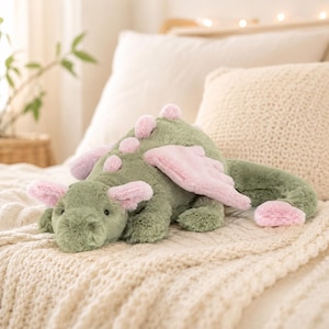 May include: A plush dragon toy featuring soft sage green fur with contrasting light pink accents on the ears, wings, tail tip, and a row of small rounded spikes running along its back. The stuffed creature has a rounded snout and dark eyes.