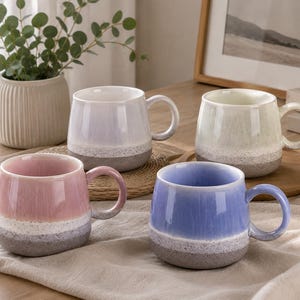 Può includere: Un set di quattro tazze in ceramica con un design a smalto reattivo bicolore. Ogni tazza presenta una parte superiore liscia e lucida in rosa pastello, lavanda, azzurro o crema, che sfuma in una base grigia testurizzata e maculata.
