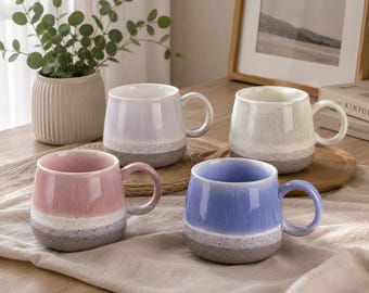 May include: A set of four ceramic mugs featuring a two-tone reactive glaze design. Each mug has a smooth, glossy upper section in pastel pink, lavender, light blue, or cream, transitioning into a textured, speckled grey base with a curved handle.