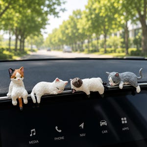 May include: Four small cat figurines perched on the edge of a car dashboard screen. The cats include a calico sitting upright, a white cat lying down, a Siamese-style cat resting, and a grey and white cat hanging over the edge of the display.