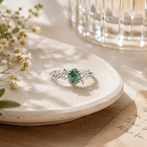 May include: A silver ring featuring a round, mottled green gemstone set in the centre. The band is designed with delicate, sculpted metal leaves and vines extending from the setting, creating a nature-inspired aesthetic for this jewellery piece.