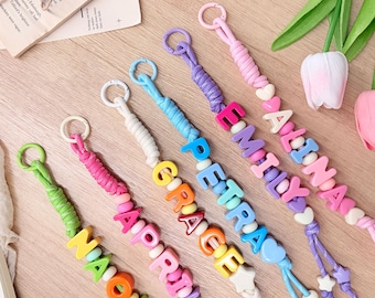 Può includere: Cinque cordini colorati con perline a lettera in plastica che compongono i nomi Naomi, April, Grace, Petra, Emily e Alina. Ogni cordino include un anello circolare, sezioni di corda avvolta, perline a forma di cuore e ciondoli a stella.