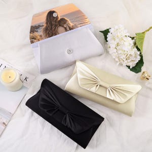 May include: Three rectangular clutch bags in black, champagne, and white satin. The black and champagne bags feature a pleated bow detail on the front flap. The white bag is open, revealing a printed photograph of two people at sunset on the interior flap.