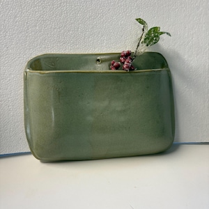 May include: A rectangular sage green ceramic wall pocket planter with rounded corners and a smooth glazed finish. A small hole is centered near the top edge for mounting. A sprig of artificial red berries with green leaves is tucked inside.