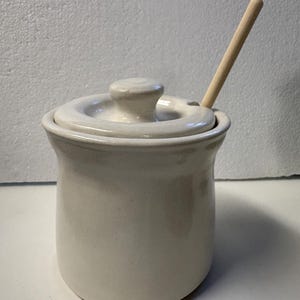 Puede incluir: Un azucarero de cerámica color crema con tapa a juego y una pequeña cuchara de madera. El cuenco tiene forma redondeada y un pequeño pomo en la tapa para facilitar su extracción. La cuchara está insertada en el cuenco.