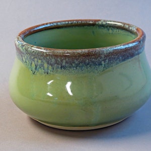 May include: A small, round ceramic bowl with a glossy finish. The bowl is primarily a light green color, with a darker green interior and a brown rim. The rim has a textured, speckled appearance. The bowl's shape is slightly wider at the base and tapers inward.