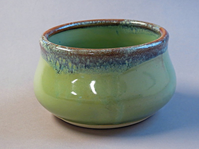 May include: A small, round ceramic bowl with a glossy finish. The bowl is primarily a light green color, with a darker green interior and a brown rim. The rim has a textured, speckled appearance. The bowl's shape is slightly wider at the base and tapers inward.