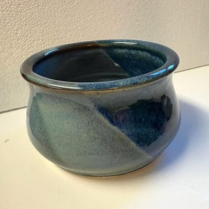 May include: A handcrafted ceramic bowl with a glossy finish. The bowl features a blend of blue and green hues, with a darker blue interior and a lighter blue-green exterior. The rim has a brown accent.