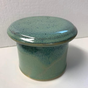 Puede incluir: Un recipiente de mantequilla de cerámica verde con tapa. El recipiente tiene un esmalte moteado y una forma ligeramente redondeada.