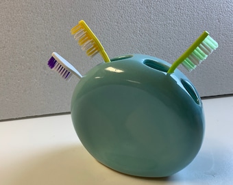 Pode incluir: Um porta-escovas de dentes de cerâmica azul claro com três ranhuras. Duas escovas de dentes estão no porta-escovas, uma com cerdas amarelas e brancas e outra com cerdas verdes e brancas. A terceira escova de dentes tem cerdas roxas e não está no porta-escovas.