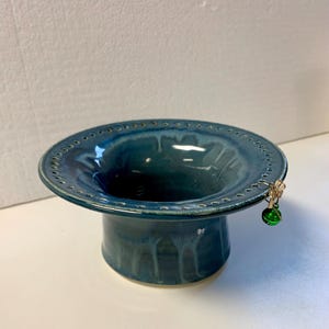 May include: A teal blue ceramic bowl with a wide rim and a small green glass ornament hanging from the side. The bowl has a textured surface and a ring of holes around the rim.