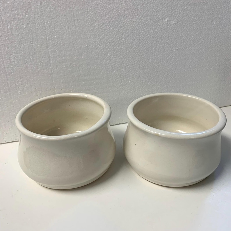 Op de afbeelding: Twee cr&egrave;mekleurige keramische kommen met afgeronde zijkanten en een brede opening. De kommen hebben een gladde, glanzende afwerking en staan naast elkaar. De binnenkant van de kommen is een lichtere tint cr&egrave;mekleurig.