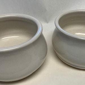 Op de afbeelding: Twee cr&egrave;mewitte keramische kommen met afgeronde lichamen en brede randen. De kommen hebben een gladde, glanzende afwerking en een eenvoudig, elegant ontwerp. Ze zijn geschikt voor serveren of decoratieve doeleinden.