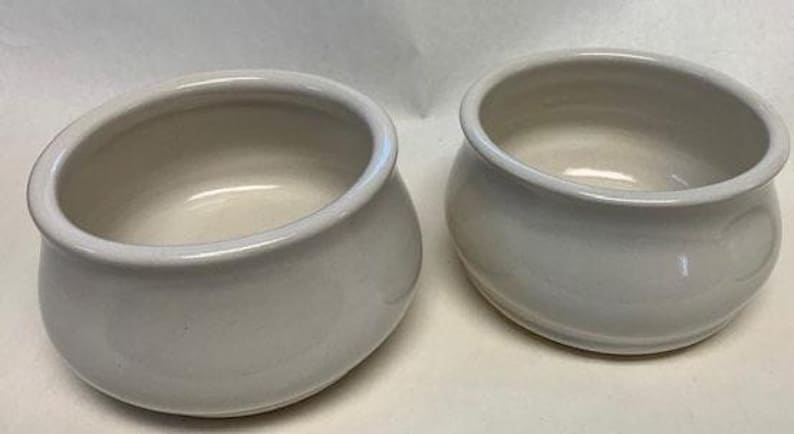Op de afbeelding: Twee cr&egrave;mewitte keramische kommen met afgeronde lichamen en brede randen. De kommen hebben een gladde, glanzende afwerking en een eenvoudig, elegant ontwerp. Ze zijn geschikt voor serveren of decoratieve doeleinden.