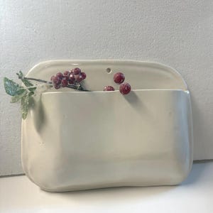 Puede incluir: Un bolsillo de pared de cerámica color crema con forma rectangular y un pequeño agujero para colgar. El bolsillo está lleno de una ramita de vegetación y varias bayas rojas, lo que sugiere un tema decorativo o de temporada.