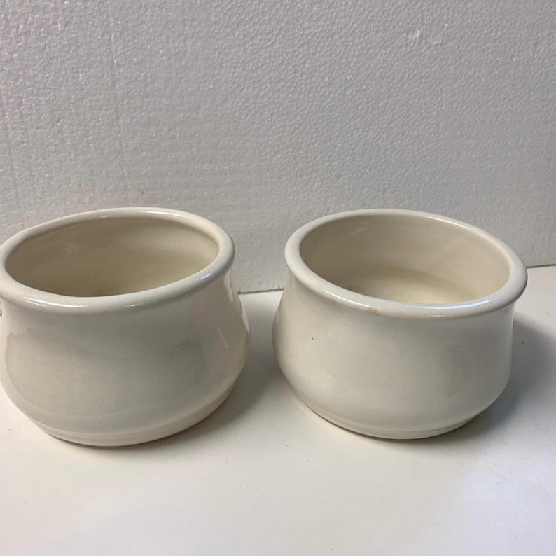 Op de afbeelding: Twee cr&egrave;mekleurige keramische kommen met afgeronde zijkanten en een licht uitlopende rand. De kommen hebben een gladde, glanzende afwerking en een eenvoudig ontwerp. De binnenkant van de kommen is lichter van kleur.