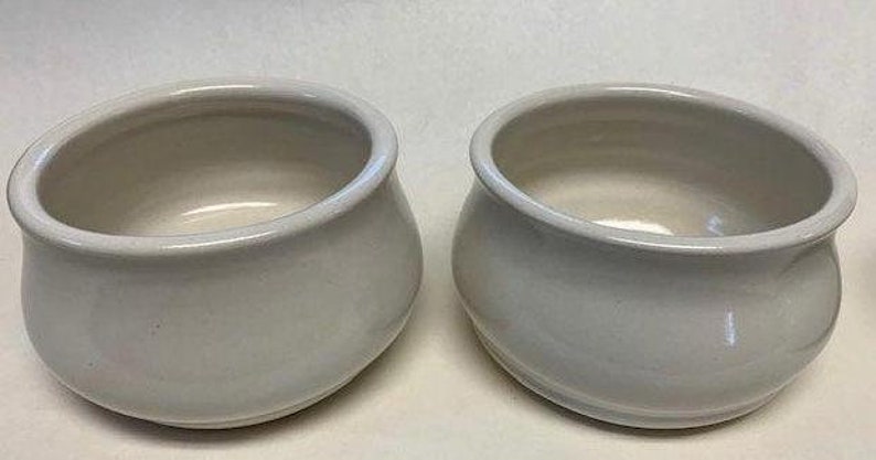 Op de afbeelding: Twee kleine, cr&egrave;mekleurige keramische kommen met afgeronde lichamen en brede, schuine randen. De kommen hebben een gladde, glanzende afwerking en een eenvoudig, elegant ontwerp. Ze zijn geschikt voor het serveren van dips, sauzen of kleine porties.