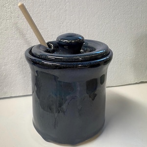 Op de afbeelding: Een donkerblauwe keramische pot met een deksel en een kleine houten lepel. De pot heeft een glanzende afwerking en een afgeronde vorm. Het deksel heeft een kleine knop bovenop. De lepel is in het deksel gestoken.