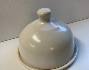 Könnte beinhalten: Eine cremefarbene Butterdose aus Keramik mit gewölbtem Deckel und kleinem Griff oben. Die Dose steht auf einem passenden kreisförmigen Sockel. Die Oberfläche hat ein gesprenkeltes Aussehen.