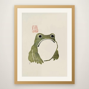 Op de afbeelding: Een ingelijste print met een gestileerde groene kikkerillustratie op een crèmekleurige achtergrond. De kikker heeft een grote witte buik, donkere ogen en een eenvoudige omtrek. Een rood vierkant zegel met Japanse tekens staat boven de kop.
