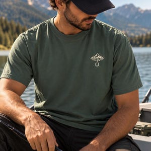 Peut inclure: Un t-shirt à manches courtes vert olive foncé avec un petit motif blanc sur la poitrine gauche. Le motif représente une chaîne de montagnes au-dessus d'un hameçon. Le t-shirt possède un col rond et une coupe standard en coton uni.