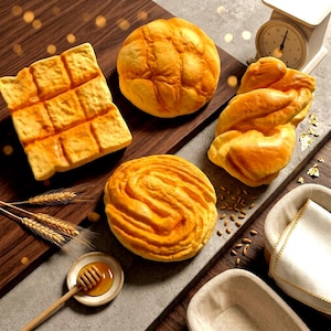 May include: A collection of four golden-brown baked bread items including a square loaf, a round bun, a twisted loaf, and a spiral roll, accompanied by a wooden honey dipper in a small bowl of honey, wheat stalks, and a white kitchen scale.