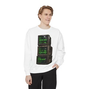 May include: A white long-sleeve sweatshirt featuring a graphic print of three stacked, weathered computer monitors. Each screen displays a cracked glass effect and the green text "SYSTEM ERROR" along with lines of technical code and data.