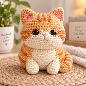 Könnte beinhalten: Ein gehäkeltes orange-weißes getigertes Katzen-Plüschtier mit großen schwarzen Kunststoffaugen und einer kleinen rosa gestickten Nase. Die Katze hat spitze Ohren mit rosa Innendetails, einen runden Körper und einen gestreiften Schwanz aus Garn.