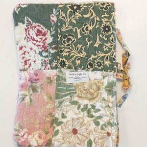 May include: A rectangular quilted fabric cover featuring a patchwork design of floral patterns in shades of sage green, dusty pink, cream, and yellow. A white fabric label in the center reads Walk in Light Co. 1 John 1:7. A fabric tie is attached.