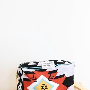 May include: A rectangular zippered pouch featuring a bold geometric pattern in black, white, red, yellow, and light blue. A white fabric label is sewn on the top edge with the text Walk in Light Co. and 1 John 1:7 printed in black ink.
