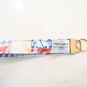 May include: A fabric wristlet keychain featuring a patchwork pattern of blue gingham, white, and red floral prints. A white rectangular tag is sewn onto the fabric with the text Walk in Light Co. and a sun icon. It has a gold metal clasp and ring.