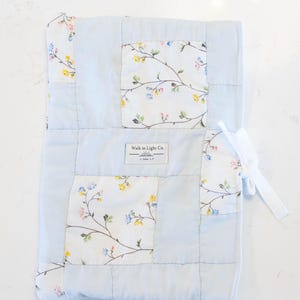 May include: A rectangular fabric cover featuring a patchwork design of solid light blue panels and white panels with a delicate floral pattern of small yellow, pink, and blue flowers. A white fabric label reads Walk in Light Co. 1 John 1:7.