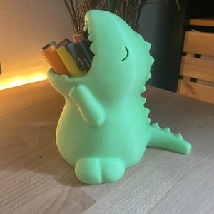 May include: A light green, 3D-printed dinosaur-shaped desk organizer with a wide-open mouth holding several colorful markers. The creature has a smooth, matte finish, a curved tail, and small arms, serving as a functional container for office supplies.