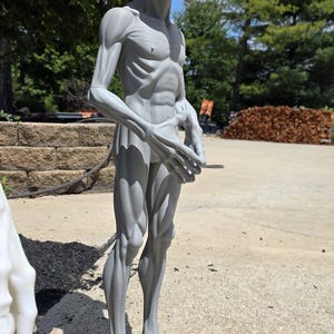 May include: A gray, standing alien figurine with a large, smooth, oval-shaped head, prominent dark eyes, and a slender, muscular build. The figure features detailed anatomical definition across the torso, arms, and legs, with long, thin fingers and feet, presented in a full-body view.