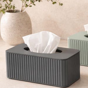 May include: A rectangular dark grey tissue box cover featuring a vertical ribbed texture on all four sides. The top surface has a smooth finish with a central oval opening, through which a white tissue is pulled upward and displayed prominently.