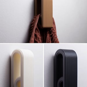 May include: A set of three wall-mounted hooks featuring a ribbed, U-shaped design. One hook is tan and holds a brown towel, while the other two are solid white and black. Each hook has a rounded top and a circular opening at the base for hanging.