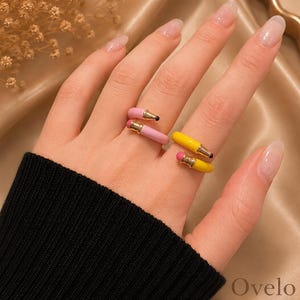 May include: A gold-toned open ring featuring two pencil-shaped designs. One pencil is pink with a gold ferrule and a black tip, while the other is yellow with a gold ferrule and a pink eraser tip. The ring is worn on a finger, showcasing the colorful enamel finish.