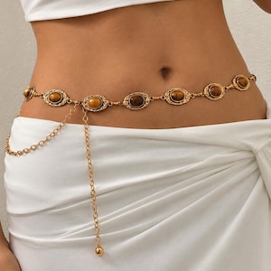 May include: A gold-toned waist chain featuring a series of oval-shaped links with embedded brown tiger's eye stones. The belt includes a delicate dangling chain segment that hangs down from the main waist band, finished with a small gold bead.