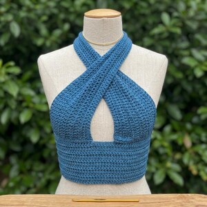 May include: A blue crochet halter top featuring a crisscross neckline design and a wide ribbed band at the waist. The garment is displayed on a mannequin, and a gold-colored crochet hook is positioned horizontally on the surface below the item.