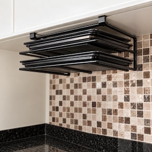 Op de afbeelding: Een zwart metalen opbergrek voor onder een kast met twee horizontale niveaus voor het opbergen van platte keukenartikelen. Het rek is bevestigd aan de onderkant van een witte kast en bevat twee rechthoekige zwarte bakplaten op de planken.