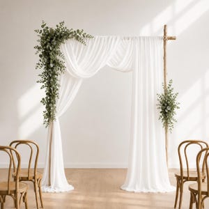 May include: A wooden wedding arch draped with sheer white fabric, featuring a large arrangement of green eucalyptus leaves on the left side and a smaller matching floral cluster on the right side, positioned between two wooden chairs.