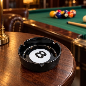 May include: A round black ceramic ashtray featuring a white circular center with a black number eight graphic. The ashtray has three notches along the rim for resting items and a glossy finish, designed to resemble a classic eight ball from billiards.
