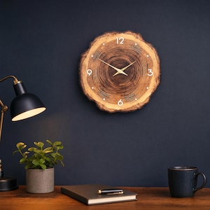 Può includere: Un orologio da parete realizzato con una fetta circolare di legno naturale con venature visibili. Il quadrante presenta numeri argentati alle posizioni 12, 3, 6 e 9, piccoli punti argentati per le altre ore e lancette in metallo dorato.