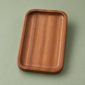 Könnte beinhalten: Ein rechteckiges Holztablett mit abgerundeten Ecken und einer glatten, polierten Oberfläche. Das Tablett weist eine warme, mittelbraune Holzmaserung mit sichtbaren natürlichen Texturen und einen erhöhten Rand um die flache Mitte auf.