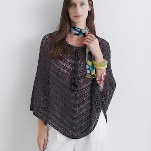May include: A dark charcoal gray knit poncho featuring an openwork chevron pattern and a drawstring neckline with tassel details. The garment is layered over a light-colored top and paired with a colorful patterned neck scarf and two lime green bangles.