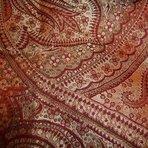 May include: Close-up of a red, gold, and brown paisley patterned fabric. The fabric has a woven texture and a repeating pattern of intricate floral designs.
