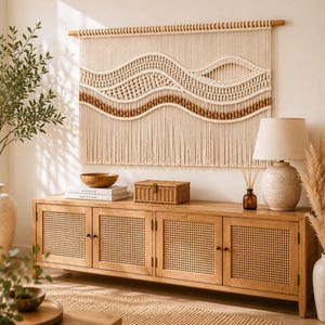 Puede incluir: Un gran tapiz de macramé color crema con patrones ondulados y detalles marrones colgado de una barra de madera. Debajo, un aparador de madera clara con cuatro puertas de rejilla, una caja pequeña tejida, un cuenco de madera y una lámpara de mesa.