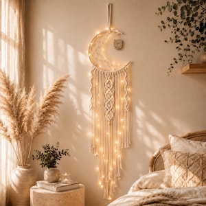 May include: A crescent moon-shaped macrame wall hanging made of off-white cotton cord, featuring intricate woven patterns and long fringe. The piece is illuminated by warm white string lights and includes a small wooden owl charm attached to the frame.