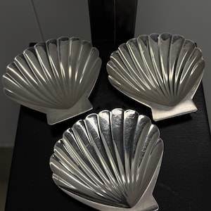 May include: Three silver-toned metal dishes shaped like scallop shells. Each dish features a fan-like design with radiating ridges extending from a flat base to a curved, scalloped edge. The metallic surfaces have a reflective finish with visible light reflections.