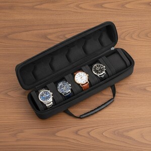 Puede incluir: Un estuche de viaje rectangular negro para relojes con cierre de cremallera y asa. El interior está forrado con espuma negra y cuenta con cuatro compartimentos, tres de los cuales contienen relojes con esferas plateadas, azules y blancas.